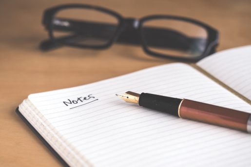 A notebook with a pen laying on top of it and glasses in the background.