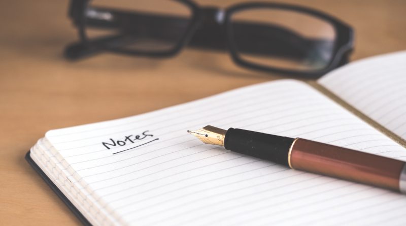 A notebook with a pen laying on top of it and glasses in the background.