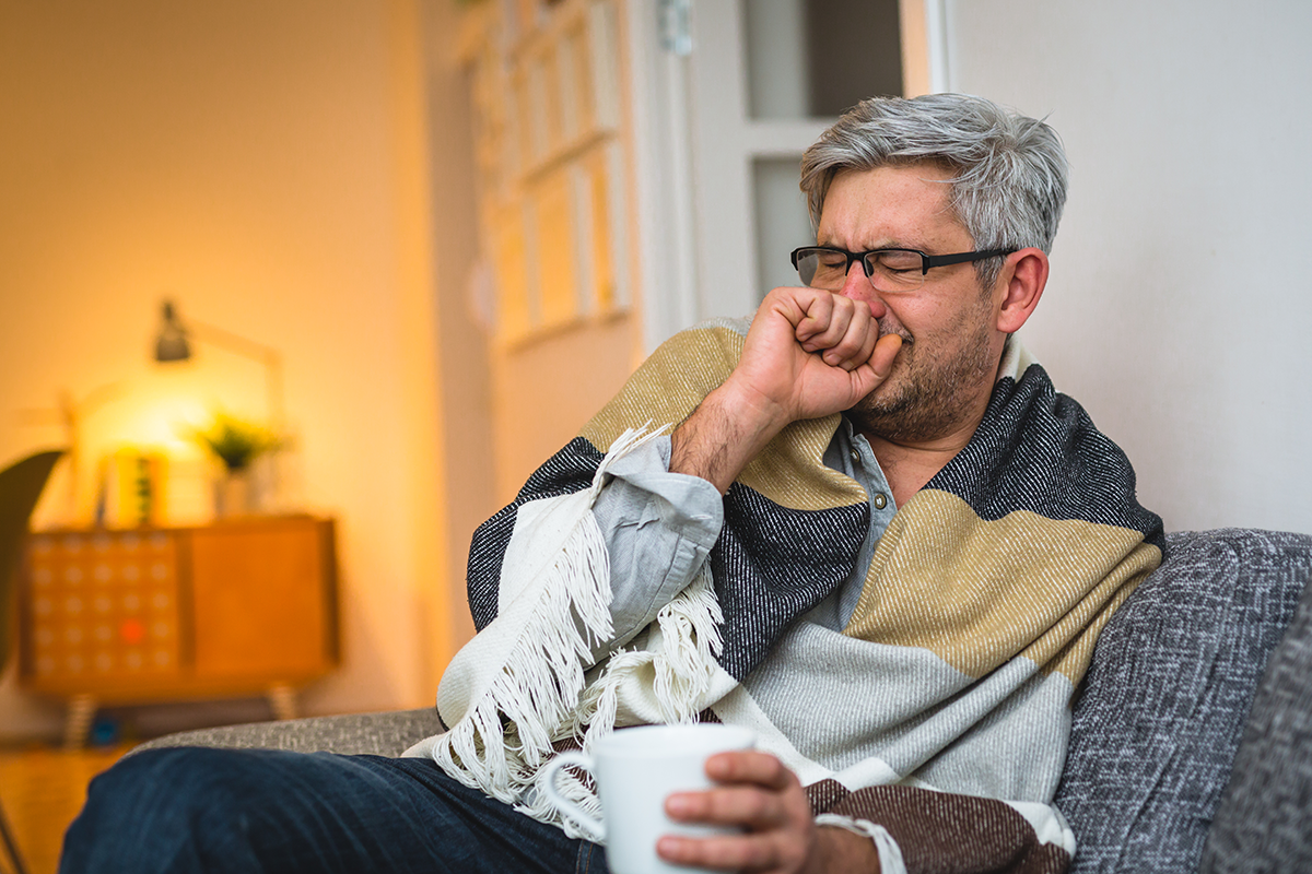 A person suffering from a cold, sitting on a couch with a blanket and a hot drink