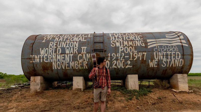 Musician Julian Saporiti stands in front of a large metal tank with the words "Justice for All" and various examples of unjust actions the American government has taken painted on it.