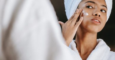 A person in a robe with their hair up in a wrapped towel looking in the mirror and rubbing moisturizer on their cheek.
