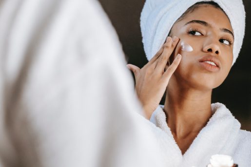 A person in a robe with their hair up in a wrapped towel looking in the mirror and rubbing moisturizer on their cheek. 