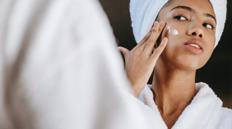 A person in a robe with their hair up in a wrapped towel looking in the mirror and rubbing moisturizer on their cheek.