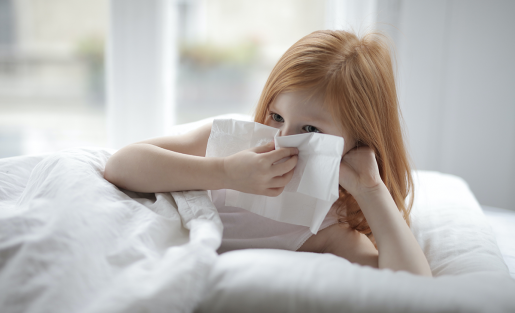 A child with a runny nose sits up in bed, blowing their nose.