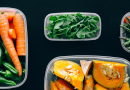 A selection of cut fruits and veggies, including leafy greens, carrots and a melon, sitting in small plastic containers on a black surface.