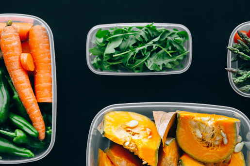 A selection of cut fruits and veggies, including leafy greens, carrots and a melon, sitting in small plastic containers on a black surface.