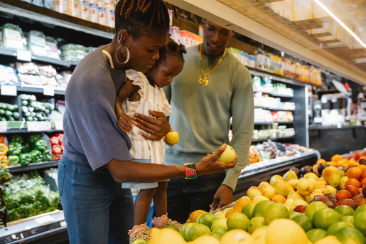 Two adults and a toddler stand together in the fruits and vegetables section of a market. The toddler is being held up by an adult, and the group is looking at apples and pears, trying to decide what to buy.
