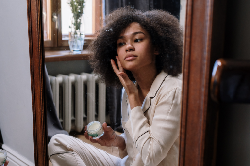 A woman seated in front of a mirror. She's applying a cream to her face to prevent wrinkles.