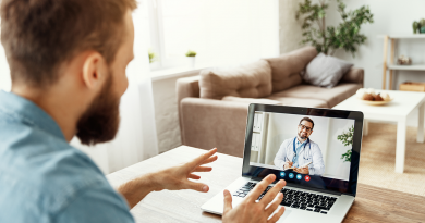 A patient sitting at their kitchen table in front of a laptop. On the laptop, a doctor discusses hernia and hernia repair via a webcam telehealth appointment.