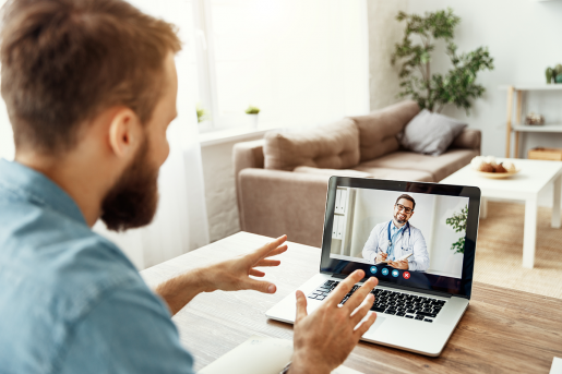 A patient sitting at their kitchen table in front of a laptop. On the laptop, a doctor discusses hernia and hernia repair via a webcam telehealth appointment.