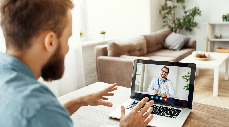 A patient sitting at their kitchen table in front of a laptop. On the laptop, a doctor discusses hernia and hernia repair via a webcam telehealth appointment.