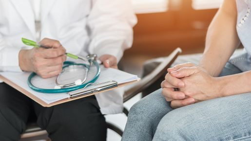 A doctor and patient seated, photographed so only their knees and mid torso are visible. The doctor is taking notes on a chart while the patient sits with arms folded.