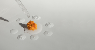 On a white and grey blank slab, a small dark-orange flower bud sits with drops of liquids around it. A dropper filled with that liquid sits above the flower bud, ready to drop more liquid on it.