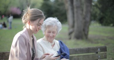 A younger person and an older person sitting together on a park bench while looking at a smart phone.