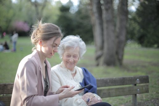 A younger person and an older person sitting together on a park bench while looking at a smart phone.