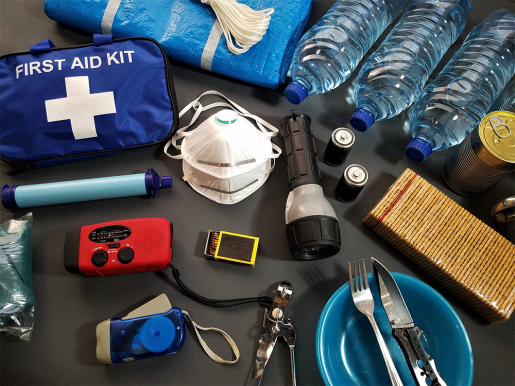 A hurricane kit, laid out on a table. Contents include silverware, a first aid kit, face masks, water bottles, batteries, a flashlight, cans of food, matches and a hand-chargeable radio.