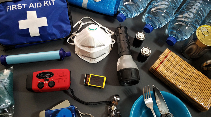 A hurricane kit, laid out on a table. Contents include silverware, a first aid kit, face masks, water bottles, batteries, a flashlight, cans of food, matches and a hand-chargeable radio.