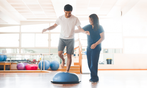 A medical professional dressed in blue scrubs offers a hand to help a patient performing a balancing exercise. The patient is balancing with one foot on top of a dome-like object.