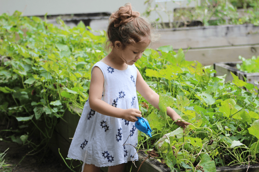 A child in a white dress holds a tool in a large garden.