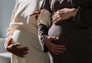 A pair of pregnant people, viewed from the side so only their torsos show. Each is holding their own stomach in an identical pose, with one hand cradling and the other resting on top. One person is wearing white, and the other is wearing dark brown. Their arms are linked.