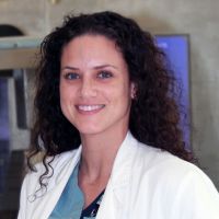 A picture of Sarah Allen. A woman with dark, curly hair dressed in a white lab coat.