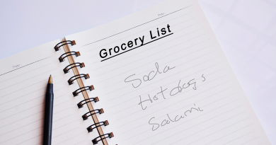 A note book sitting on a table. A hand-written grocery list shows a need to purchase soda, hot dogs and salami. A pen rests next to the list.