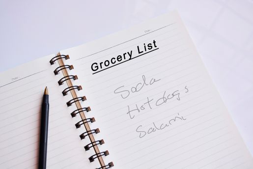 A note book sitting on a table. A hand-written grocery list shows a need to purchase soda, hot dogs and salami. A pen rests next to the list.