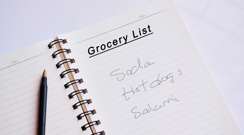 A note book sitting on a table. A hand-written grocery list shows a need to purchase soda, hot dogs and salami. A pen rests next to the list.