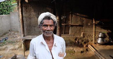 An older Indian man in a villaige in the mountains of Munnar, India. He stands wearing a white shirtwith most of the buttons undone and a white turban. His short beard is white and his hair is turning a bit grey.