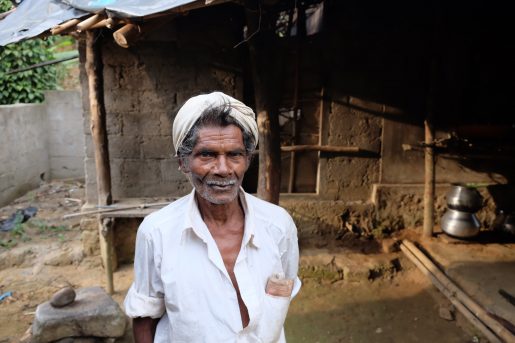 An older Indian man in a villaige in the mountains of Munnar, India. He stands wearing a white shirtwith most of the buttons undone and a white turban. His short beard is white and his hair is turning a bit grey.