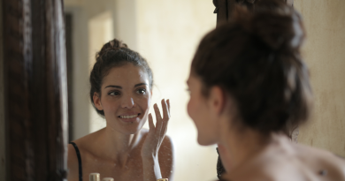 A person with dark hair pulled up into a bun is looking into a mirror while applying a skin cream.