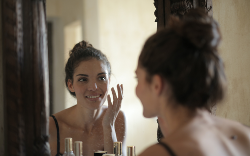 A person with dark hair pulled up into a bun is looking into a mirror while applying a skin cream.