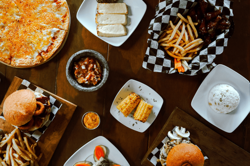 A collection of food, such as burgers and fries, pizza, chicken sings, roasted corn and rice. The food is placed on a table and shot from above.