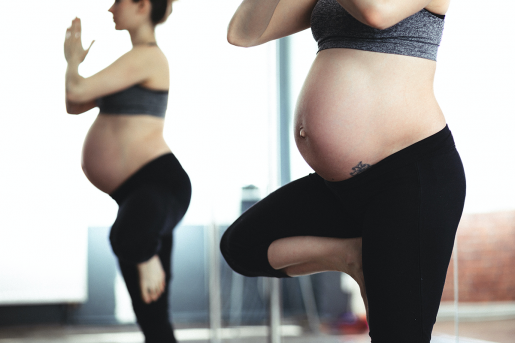 A pregnant person with a large, exposed stomach stands in a yoga pose - hands clasped at shoulder height, one leg tucked into the other.