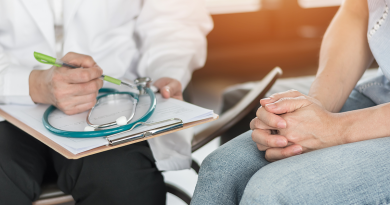 A doctor holding a clipboard seated next to a patient. Both are viewed from the neck down.