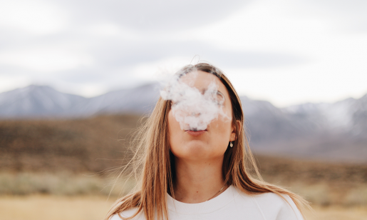 A person outdoors, standing with a mountain range visible in the distance. The person is blowing a thick cloud of vape smoke.