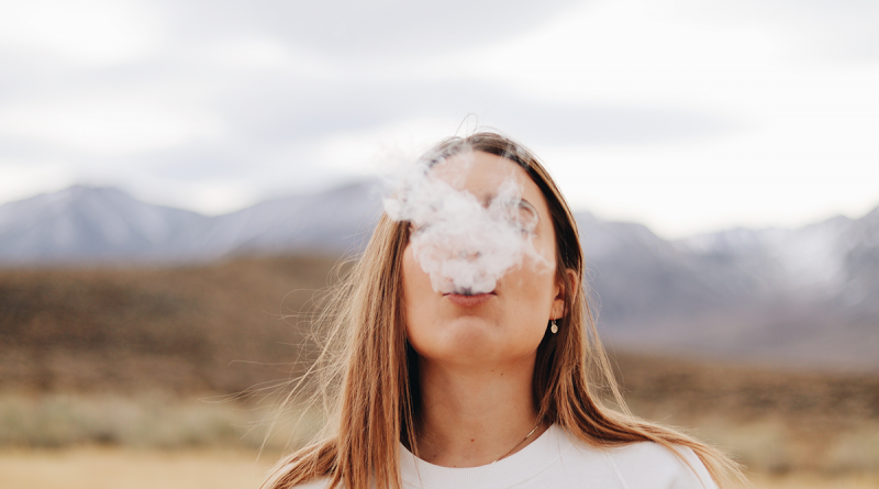 A person outdoors, standing with a mountain range visible in the distance. The person is blowing a thick cloud of vape smoke.