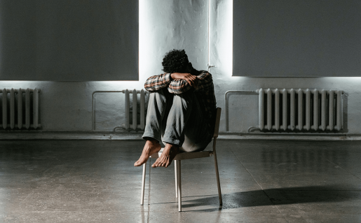 A youth with bare feet seated on a chair in an empty room with their head tucked into their knees.