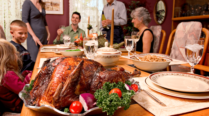 A holiday feast - turkey, dressing and vegetables. A family, including older people and children, sit around the table smiling.