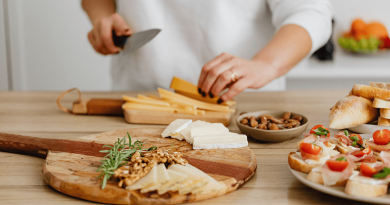 A person holding a knife and chopping slices of cheese to go along with nuts, breads and other finger foods already placed on serving platters.