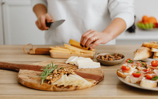 A person holding a knife and chopping slices of cheese to go along with nuts, breads and other finger foods already placed on serving platters.