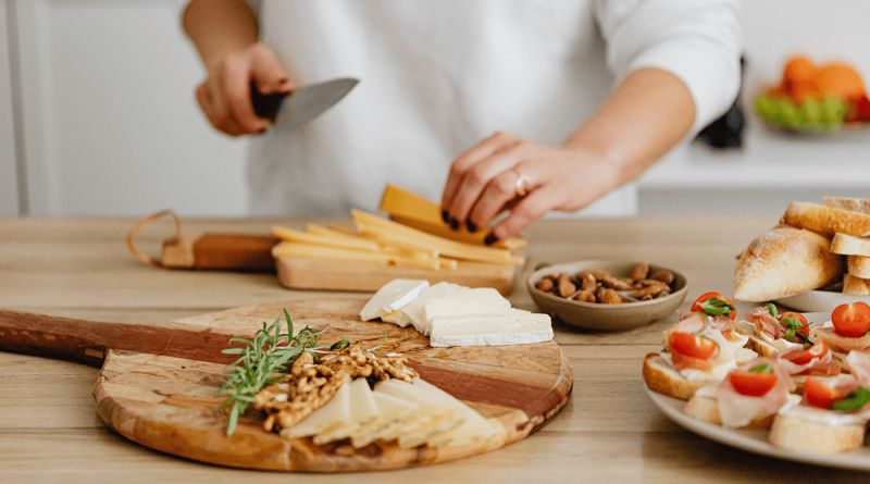 A person holding a knife and chopping slices of cheese to go along with nuts, breads and other finger foods already placed on serving platters.
