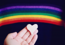 A small white heart, held in a hand in front of a painted rainbow.