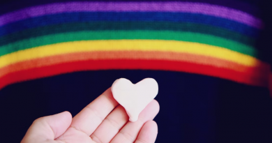 A small white heart, held in a hand in front of a painted rainbow.