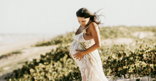 A pregnant woman in white holding her large baby bump while walking on a beach.