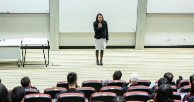 A female teacher standing at the front of a lecture hall speaking to students.