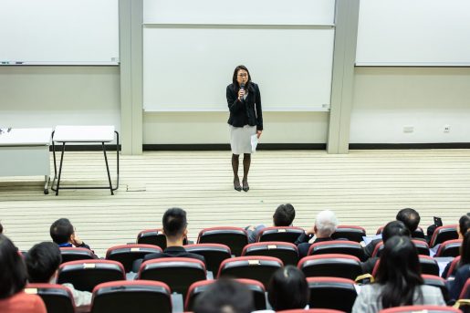 A female teacher standing at the front of a lecture hall speaking to students.