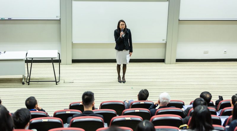 A female teacher standing at the front of a lecture hall speaking to students.