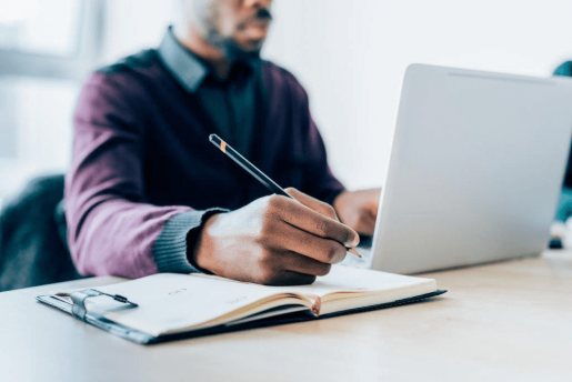 A person writing in a notebook while working on a laptop.