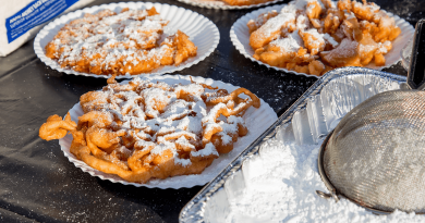 A trio of fried funnel cakes dusted with powder sugar sit on a picnic table next to a powdered sugar sifter.